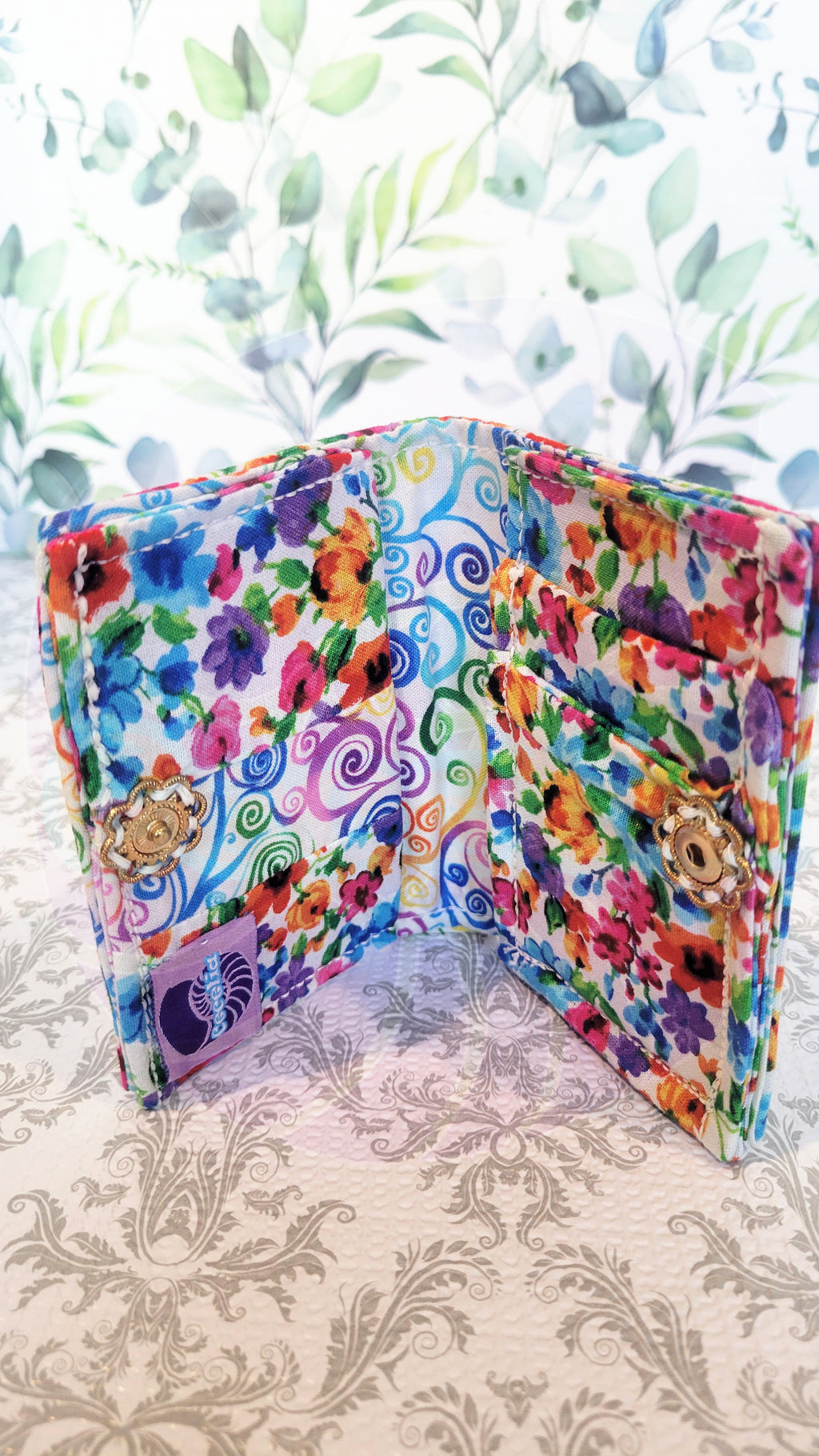 Colorful floral wallet on a patterned surface. Open to show four pocket one side of the white wallet and one pocket on the other side of the wallet. In the middle it shows colorful swirls on white