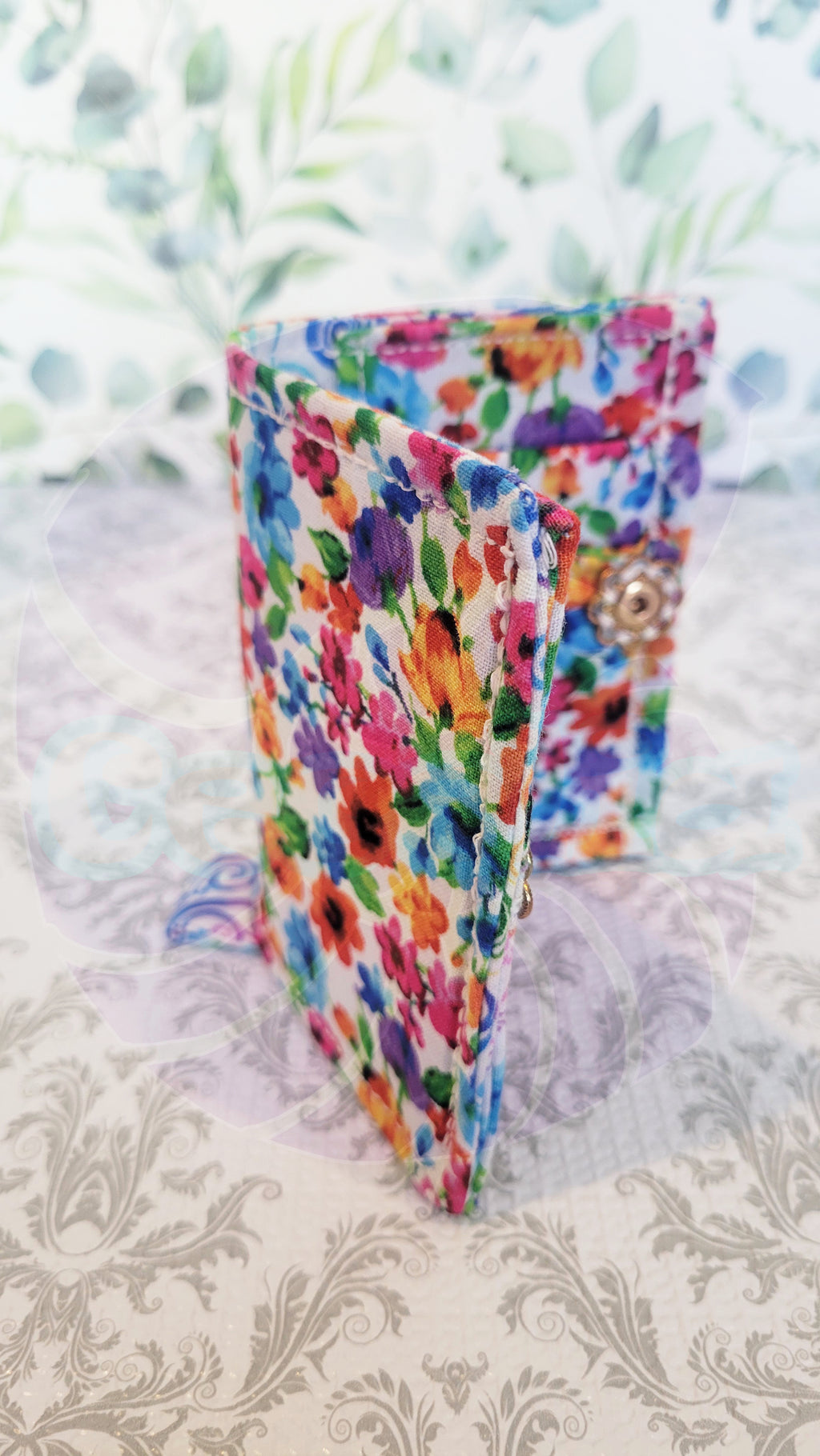Colorful floral wallet on a patterned surface. It is open in book position and a gold vintage flower push button is sewing on with white thread.