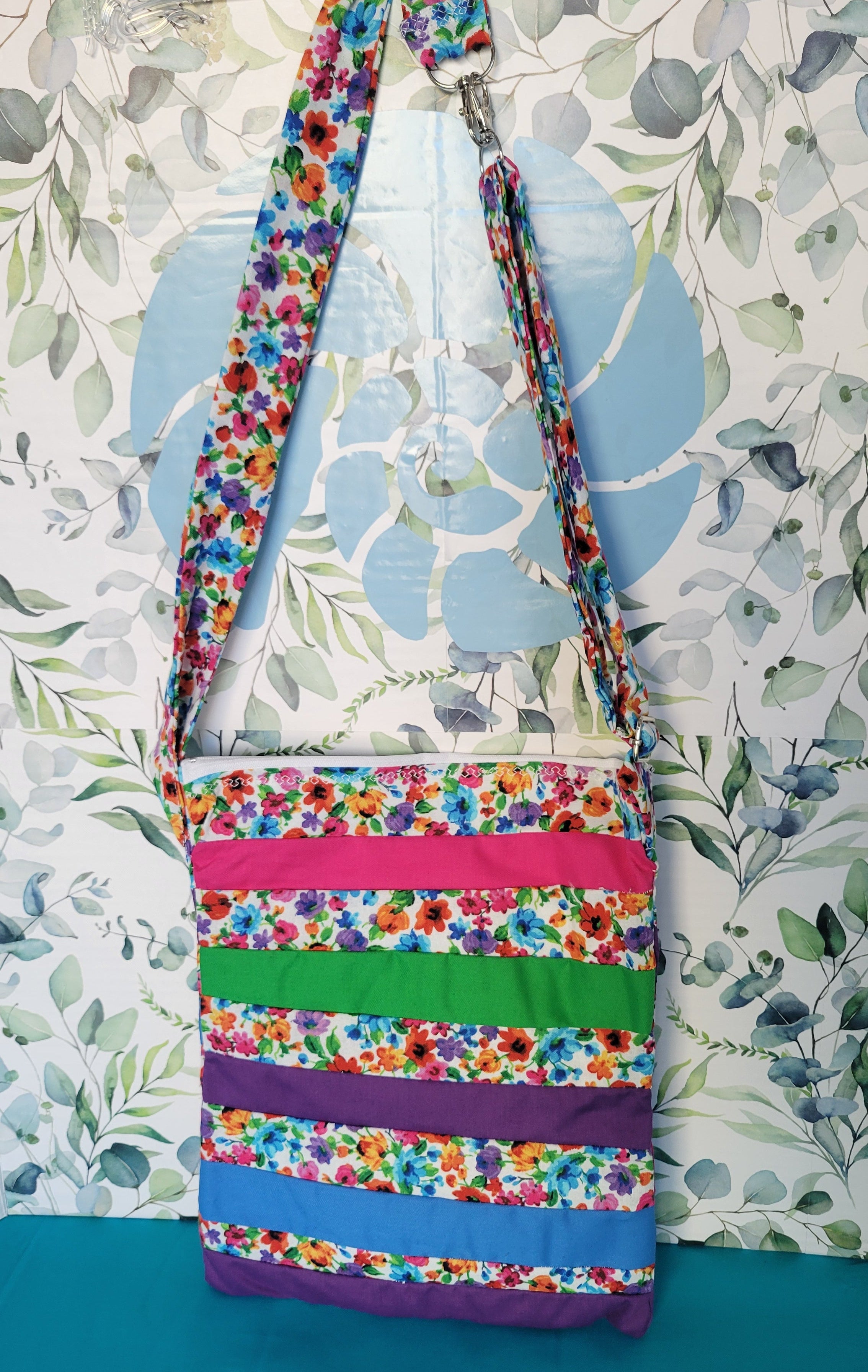Colorful floral-patterned bag hanging on its adjustable strap in the same pattern as the purse. The purse is made of strips of the colorful floral patterned and in between the strips are strip with the colors of green, purple, blue, and pink. The Purse is closed.