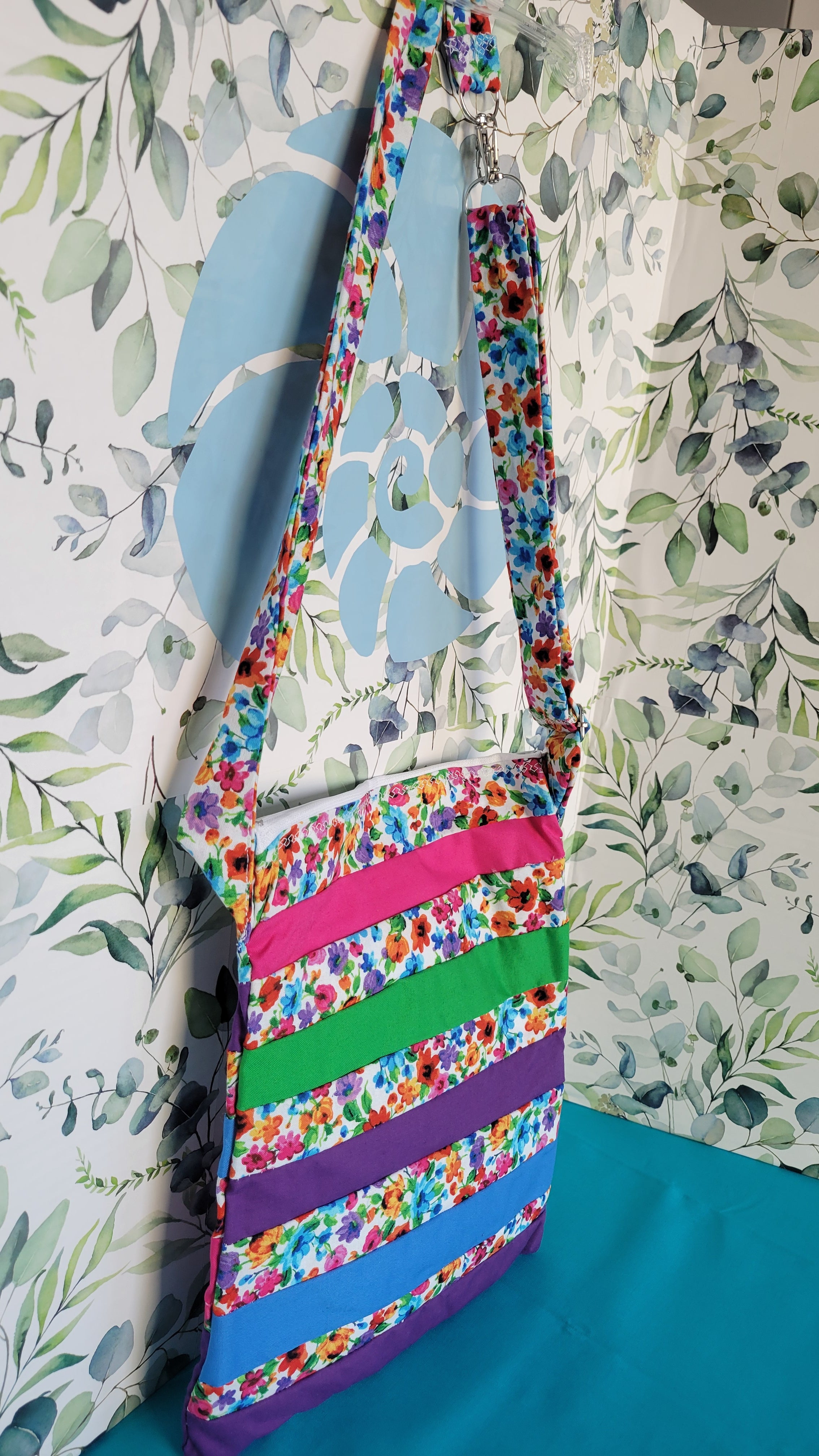 Colorful floral-patterned bag hanging on its adjustable strap in the same pattern as the purse. The purse is made of strips of the colorful floral patterned and in between the strips are strip with the colors of green, purple, blue, and pink. It is open to show its one pocket inside the purse. The inside is all white.
