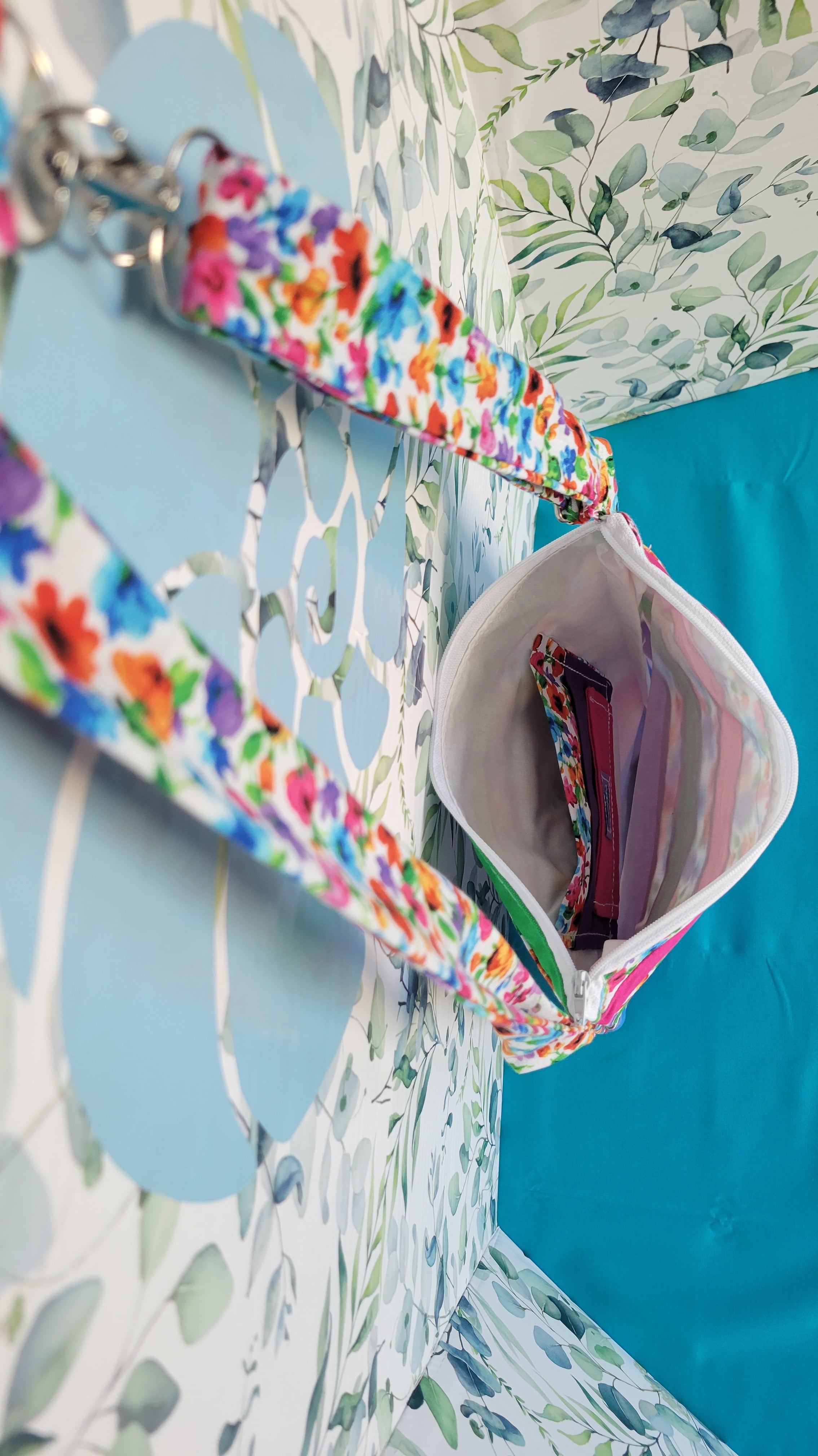 Colorful floral-patterned bag hanging on its adjustable strap in the same pattern as the purse. The purse is made of strips of the colorful floral patterned and in between the strips are strip with the colors of green, purple, blue, and pink. It is open to show its one pocket inside the purse. The inside is all white.