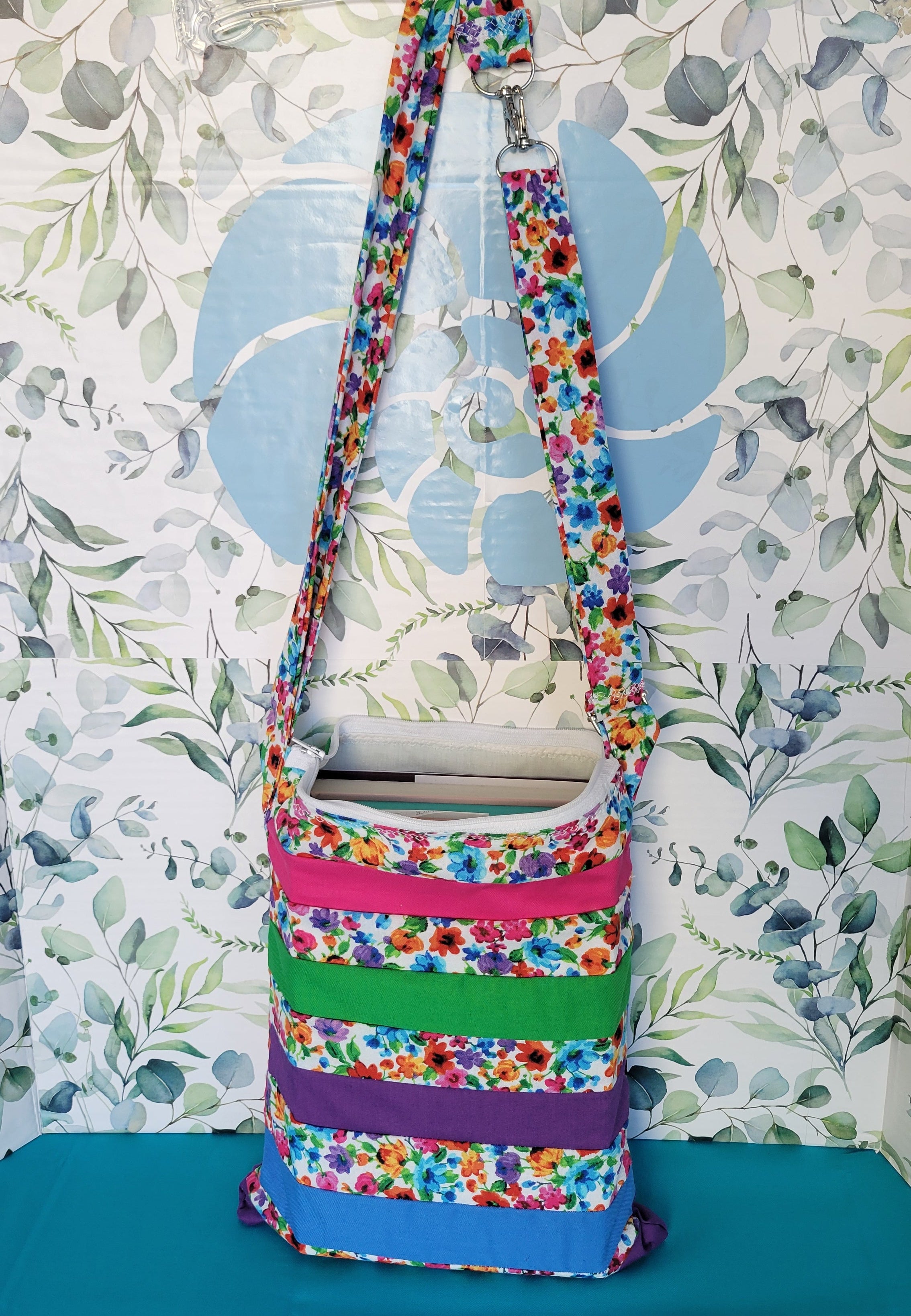 Colorful floral-patterned bag hanging on its adjustable strap in the same pattern as the purse. The purse is made of strips of the colorful floral patterned and in between the strips are strip with the colors of green, purple, blue, and pink. It is open to show a hardcover book can fit inside the purse.