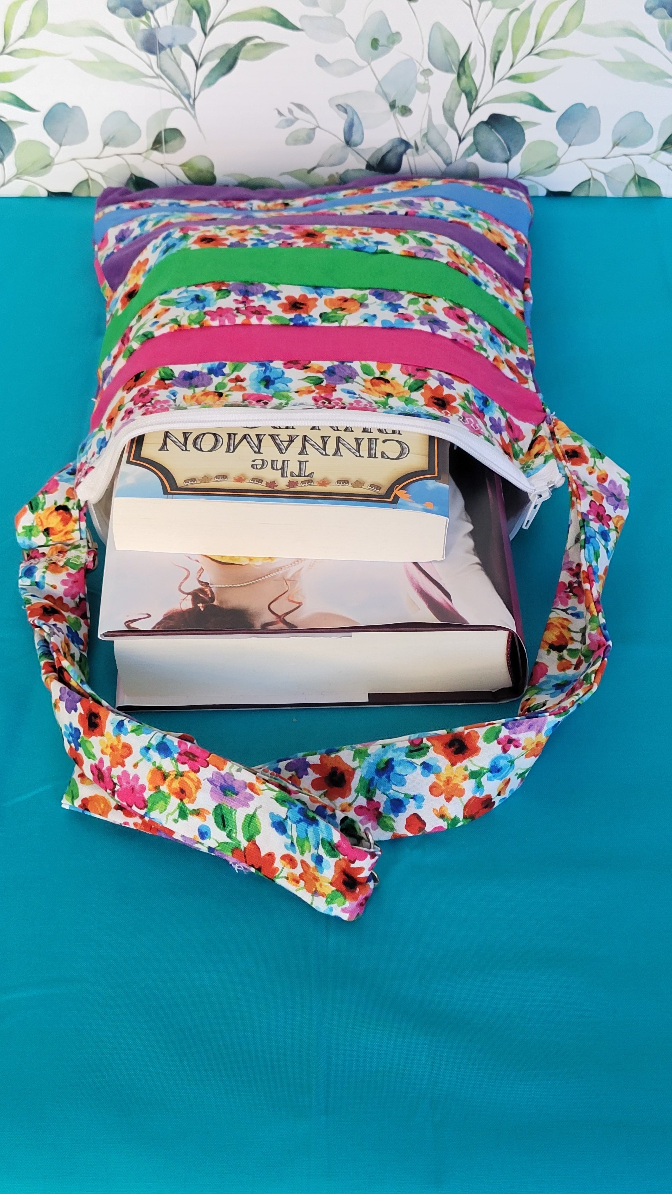 Colorful floral-patterned bag with books inside on a teal background. The purse is made of strips of the colorful floral patterned and in between the strips are strip with the colors of green, purple, blue, and pink. The purse has the same pattern adjustable strap. It is showing one soft cover and hard covering fitting inside the purse.