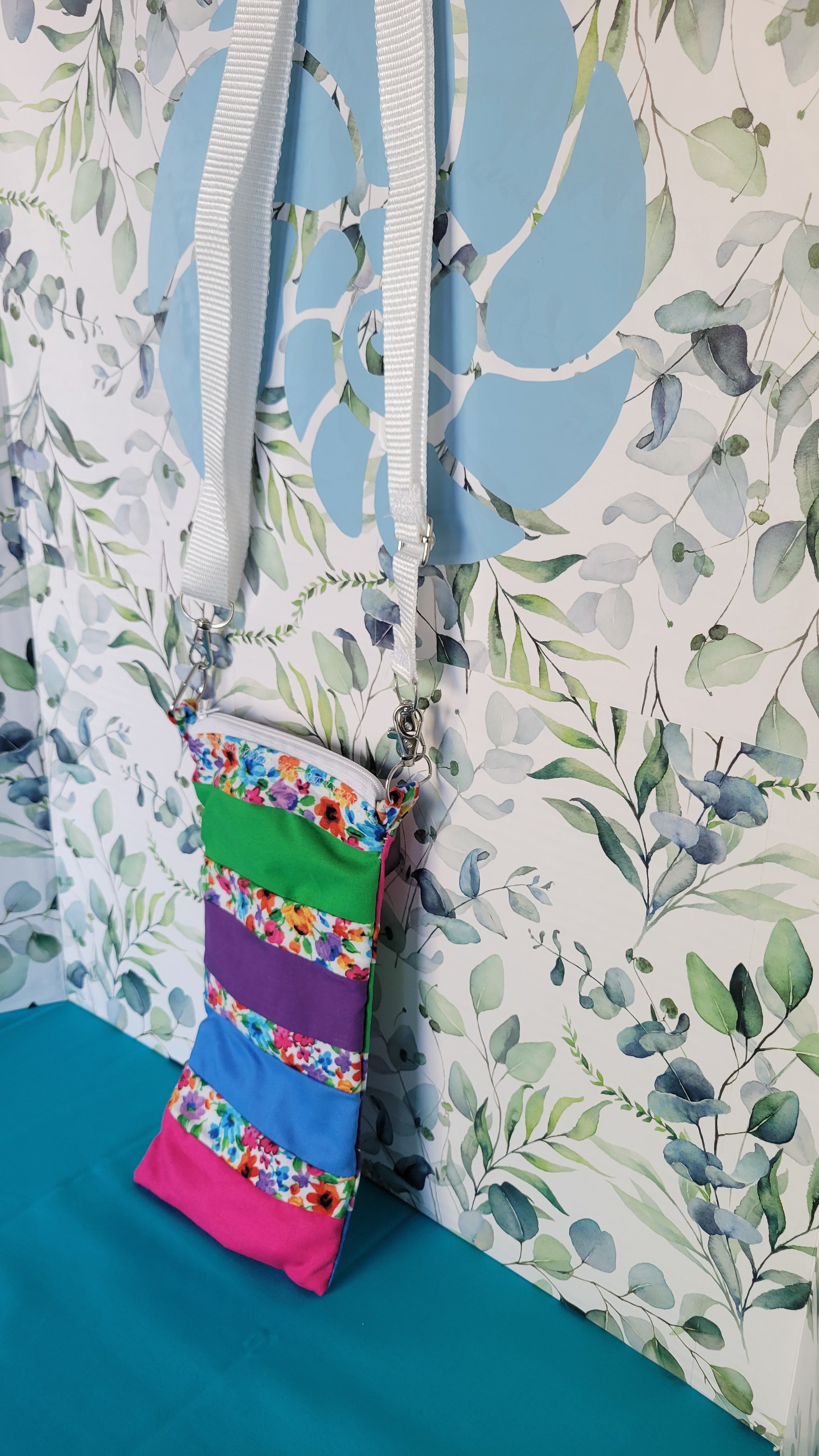 Colorful phone purse with a floral pattern. The pattern is in strips and in between the strip colors of green, purple, blue, and pink. The purse is hanging from its adjustable white strap. Phone is from the side.