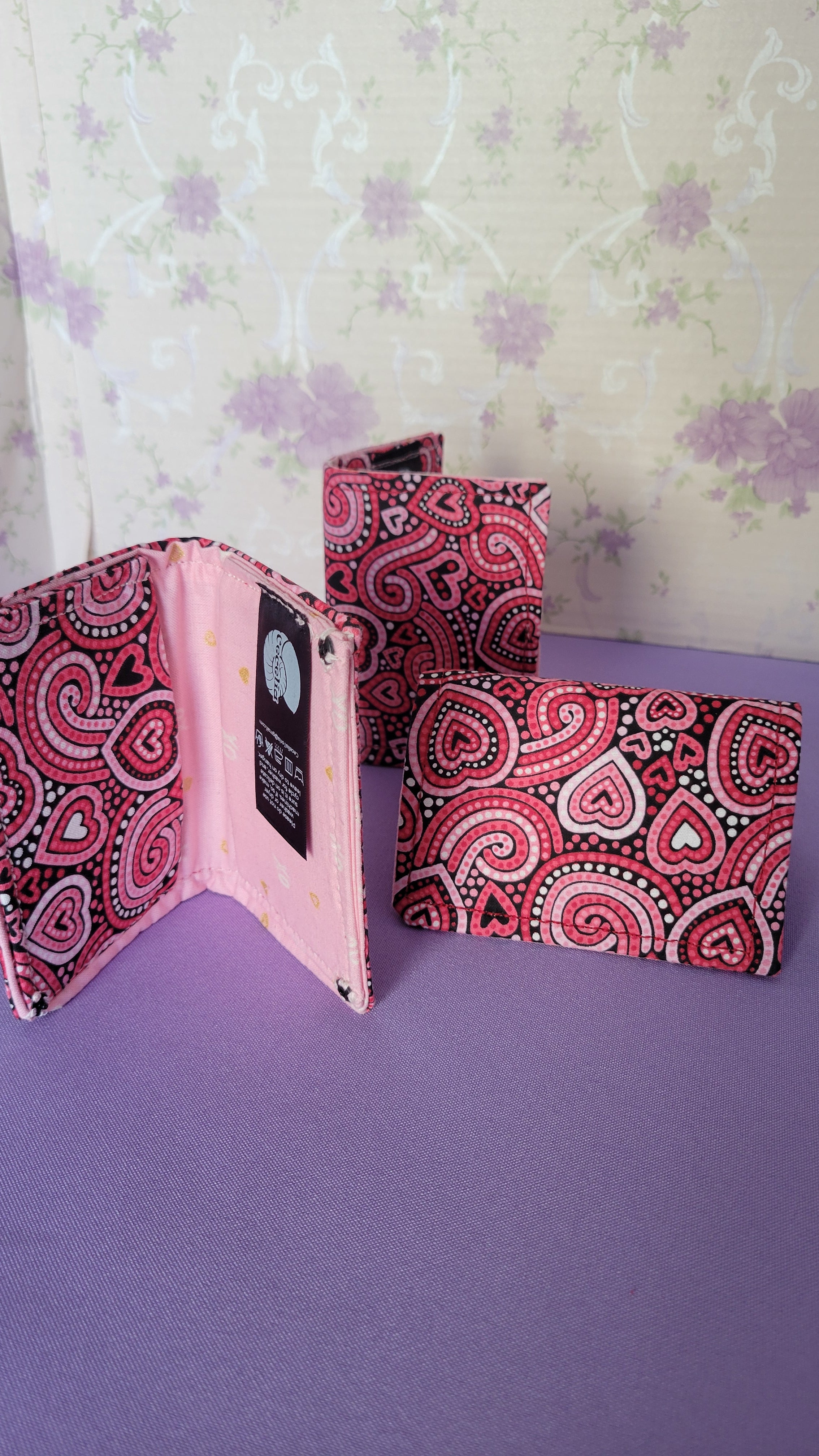 Three Javier wallets with a ripple effect of hearts in a selection of colors pink, white, red and black. They are placed parallel with one on a turning table.