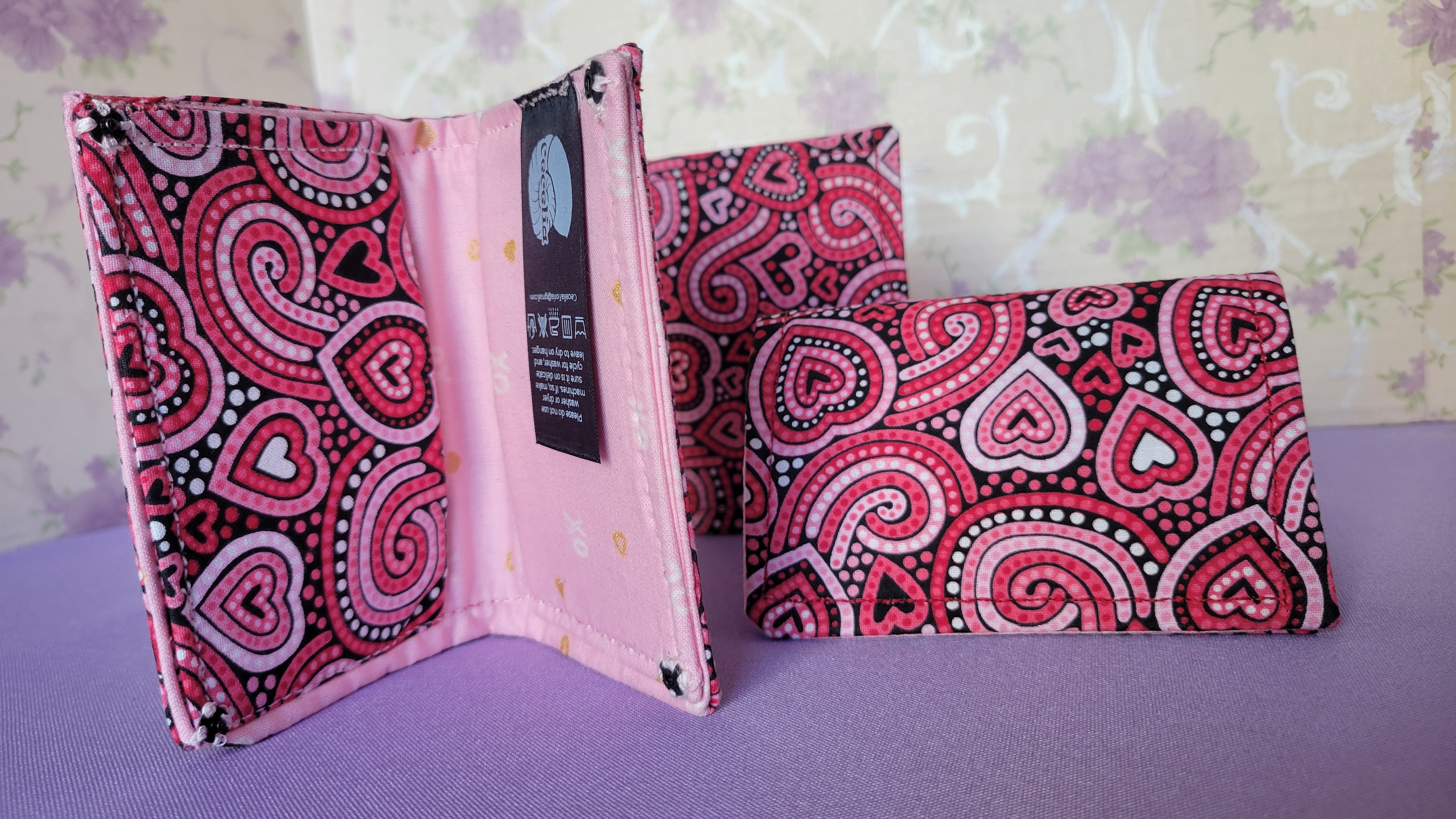 Three Javier wallets with a ripple effect of hearts in a selection of colors pink, white, red and black. They are placed parallel with one on a turning table.