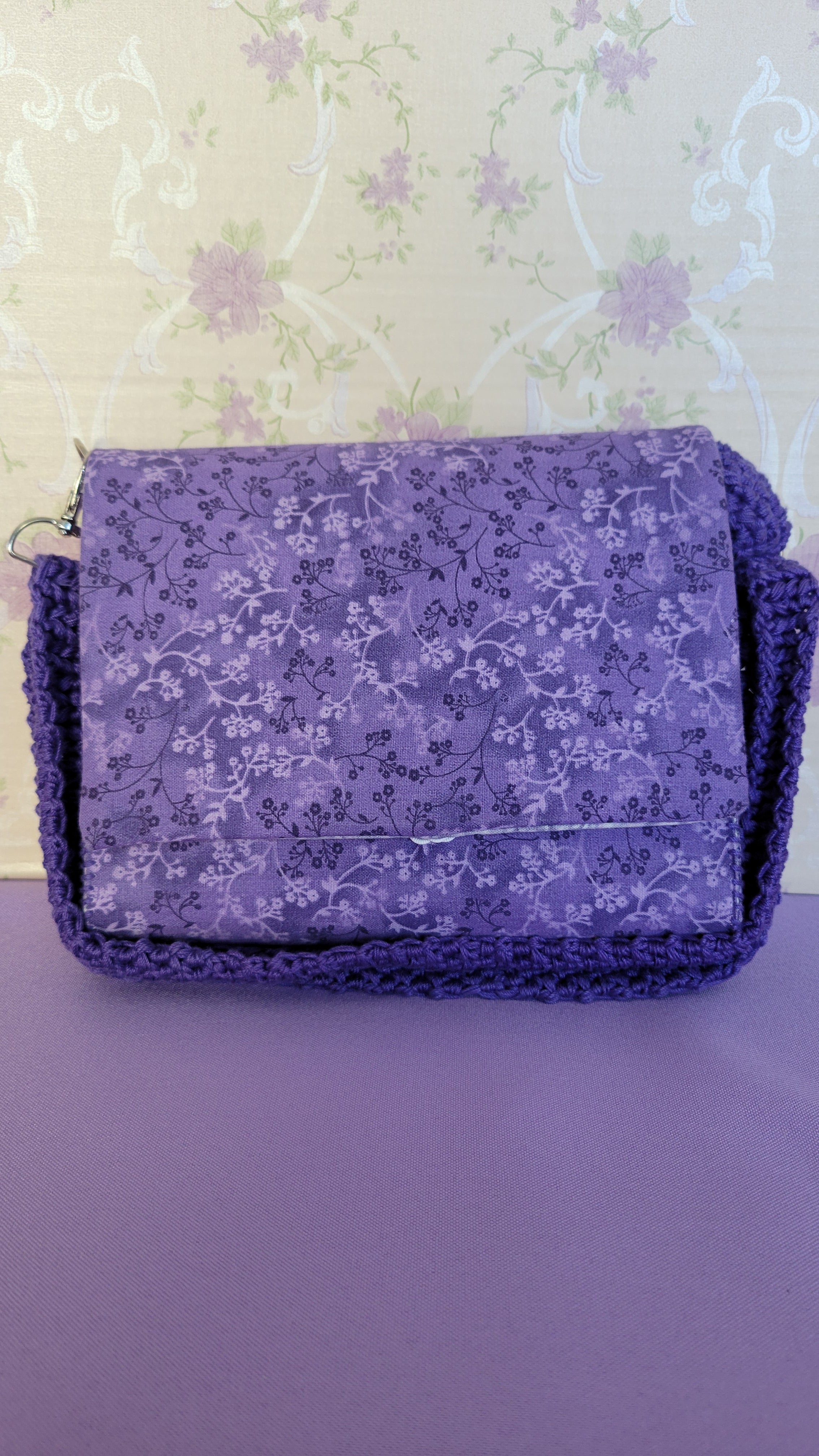 Purple clutch flores purse bag with floral patterns on a purple fabric. The Purse is leaning on the wall of a floral background in tan.