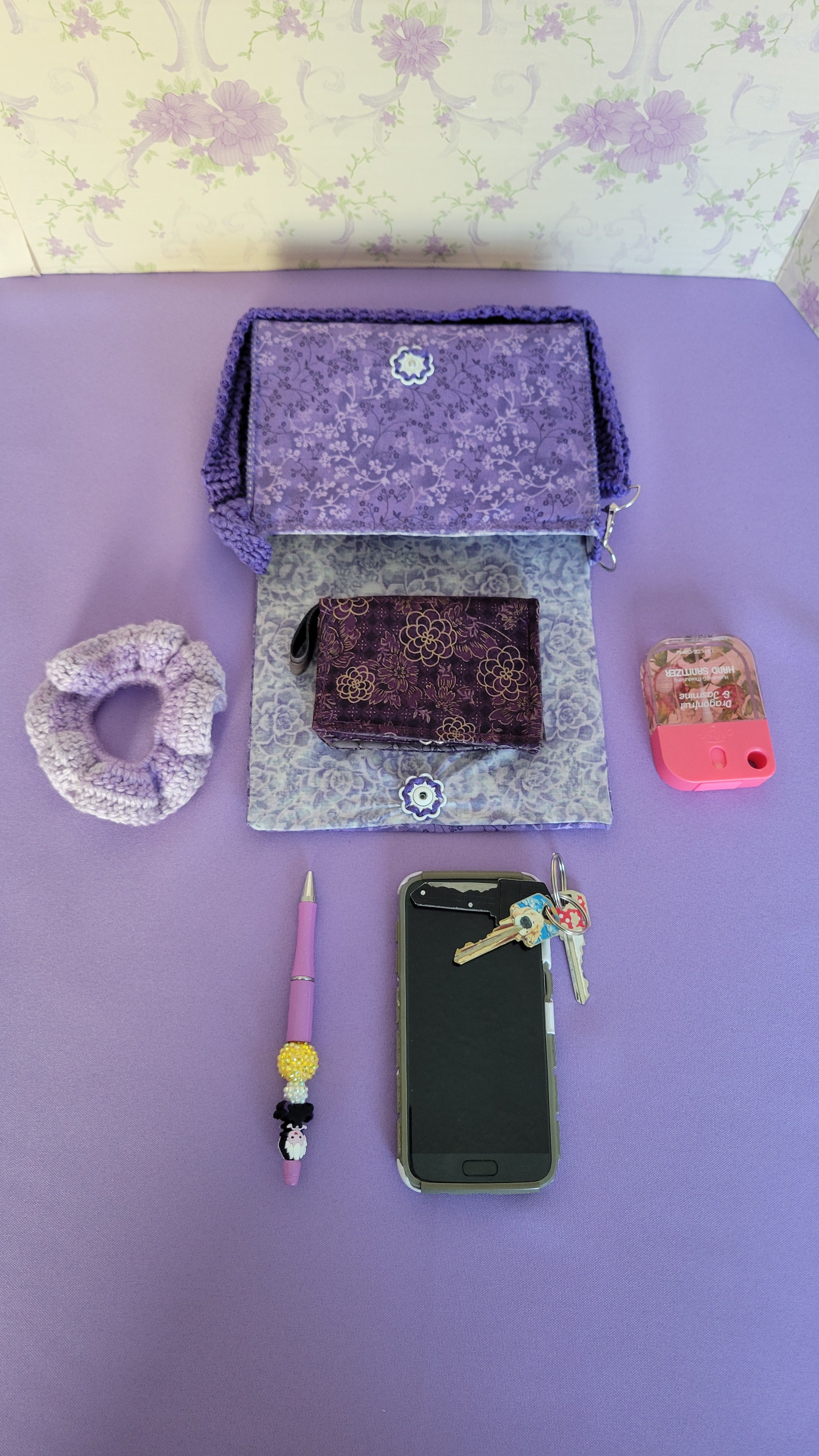 Purple clutch flores purse bag with floral patterns on a purple fabric. The purse is open showing a lavender design of roses, and a white vintage flower push bottom.
In front of the purse shows all the items it can fit. A wallet, car keys, scrunchies, and more.