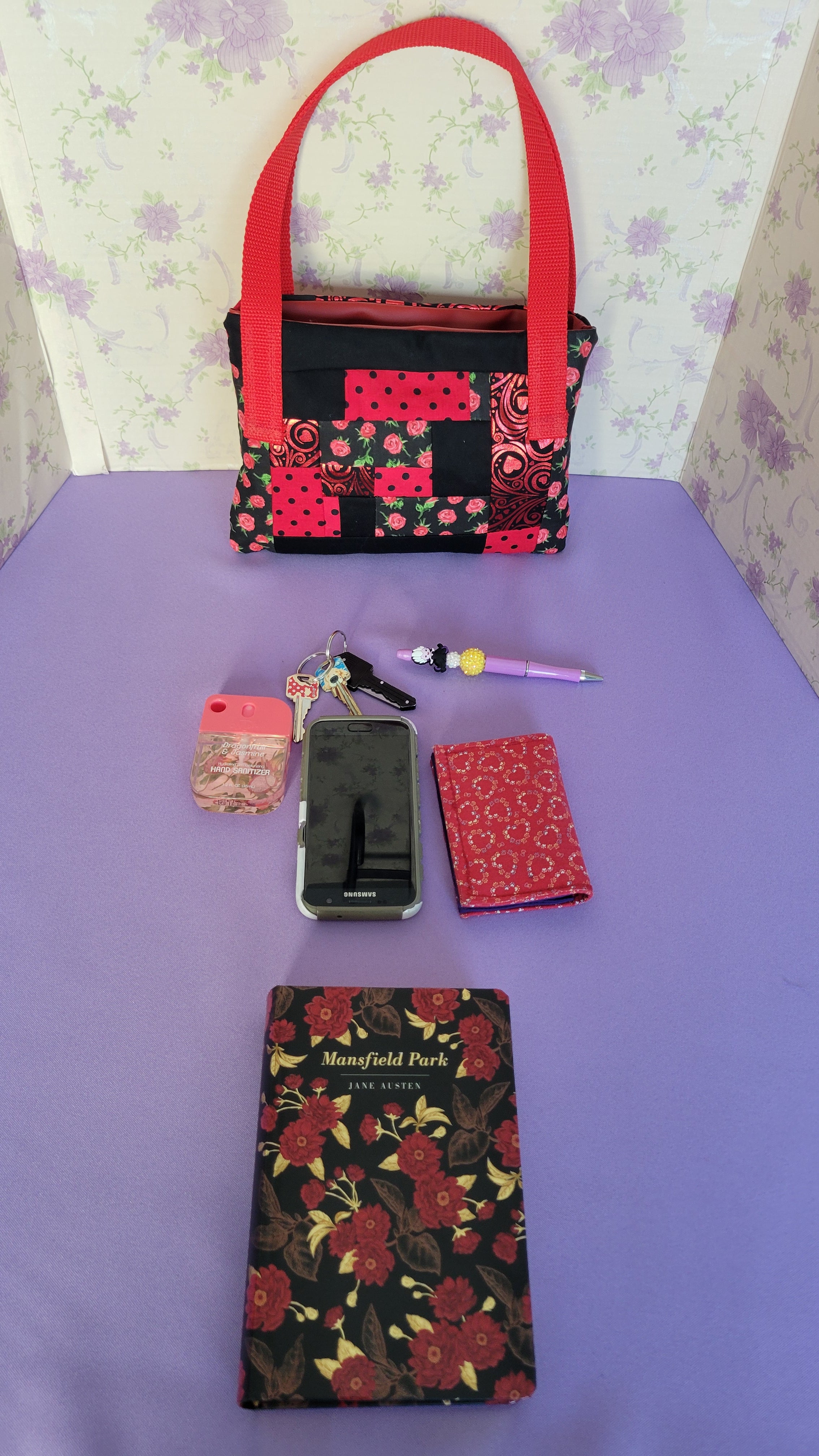 A Red Toe bag called Tracy style with two straps in red. The 5 quilt style bag has the color black., red with black polka dots, red swirls and hearts in black background, and red roses with green leaves pattern on background fabric. 

In front of the purse is items it can fit. A hand sanitizer, a red heart wallet, an example book it can fit, a touch phone and Ursula pen.